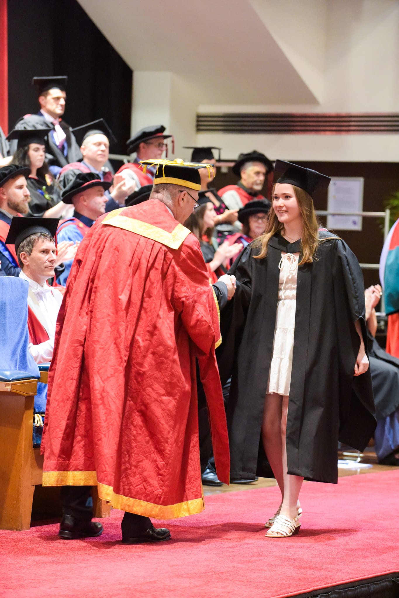 Emma, a smiling brown-haired girl in a white dress and black robes, walking across the stage and shaking hands with a man at her graduation ceremony